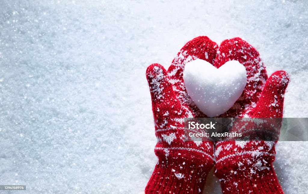 Female hands in knitted mittens with snowy heart against snow background Female hands in knitted mittens with snowy heart against snow background. Love, winter and Valentines day romantic creative concept with copy space for text Winter Stock Photo