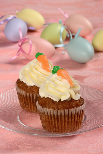 Festive carrot cake cupcakes for Easter dessert with cream cheese frosting and pastel Easter eggs in the background.