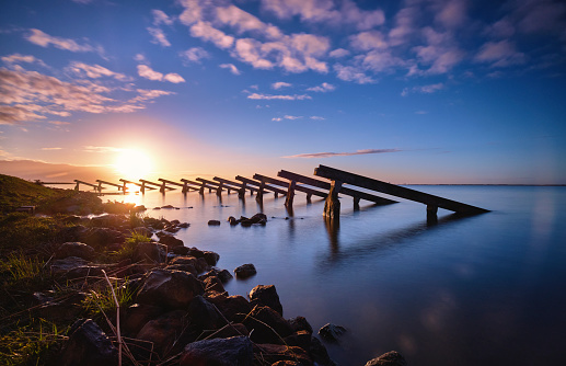 Icebreakers on the shore of Marken during sunrise. The sky is filled with clouds that are slightly pink/purple colored. The sun shines brightly in the background. The water reflects the icebreakers and the different colors of the sky. You see a part of the shore which is covered in grass and rocks.