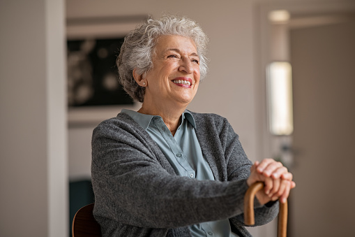 Retired senior woman laughing with her wooden walking stick while relaxing at home. Happy smiling old woman holding walking cane and looking through the window with positivity. Carefree smiling grandmother sitting on chair and looking away in nursing home.