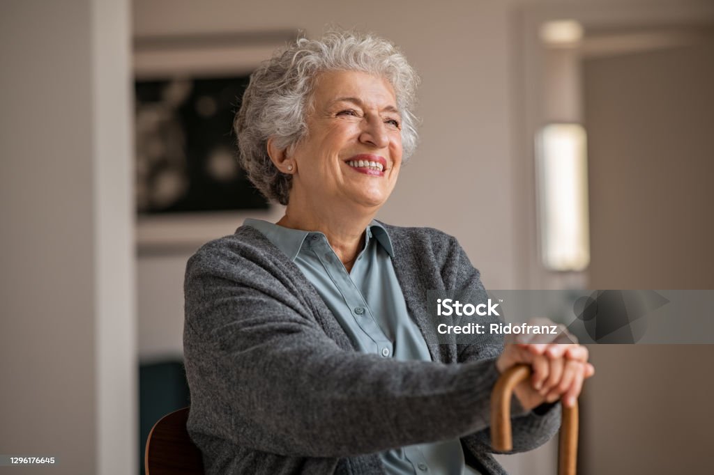 Happy smiling senior woman relaxing at home Retired senior woman laughing with her wooden walking stick while relaxing at home. Happy smiling old woman holding walking cane and looking through the window with positivity. Carefree smiling grandmother sitting on chair and looking away in nursing home. Senior Adult Stock Photo