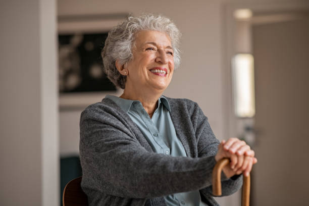 Happy smiling senior woman relaxing at home Retired senior woman laughing with her wooden walking stick while relaxing at home. Happy smiling old woman holding walking cane and looking through the window with positivity. Carefree smiling grandmother sitting on chair and looking away in nursing home. senior home stock pictures, royalty-free photos & images