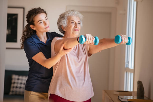 Senior woman using dumbbells with physiotherapist Old woman training with physiotherapist using dumbbells at home. Therapist assisting senior woman with exercises in nursing home. Elderly patient using dumbbells with outstreched arms in a physical therapy session in clinic. physical therapy stock pictures, royalty-free photos & images