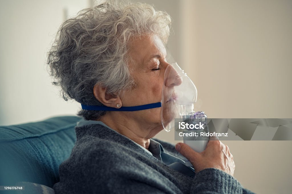 Sick senior woman making inhalation with nebulizer Portrait of a ill senior woman making inhalation at home. Close up of an elderly woman holding mask nebulizer inhaling fumes medication into lungs with closed eyes. Self treatment of the respiratory tract using inhalation nebulizer. Oxygen Stock Photo