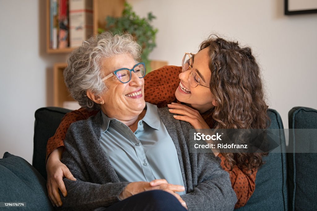 Grandmother and granddaughter laughing and embracing at home Old grandmother and adult granddaughter hugging at home and looking at each other. Happy senior mother and young daughter embracing with love on sofa. Happy young woman hugging from behind grandma with love. Senior Adult Stock Photo