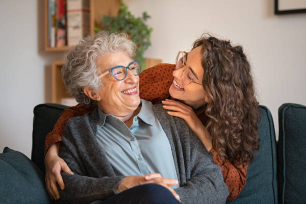 Grandmother and granddaughter laughing and embracing at home Old grandmother and adult granddaughter hugging at home and looking at each other. Happy senior mother and young daughter embracing with love on sofa. Happy young woman hugging from behind grandma with love. eyewear photos stock pictures, royalty-free photos & images