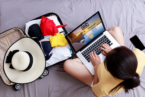 Top view Asian happy woman plan for trip by laptop booking hotel on the bed at home. Women with travel bag use notebook computer internet booking hotel. Holiday summer vacation concept