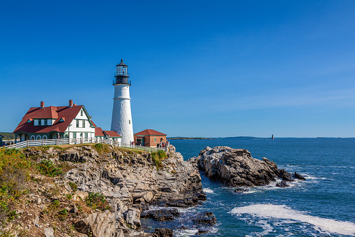 Portland Head Lighthouse, Cape Elizabeth, Maine, lit by the Morning Sun. Rocky Coastline with Cliffs, Ocean Surf and Blue Sky are in the image.