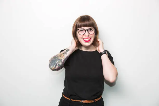 Studio portrait of a hipster nerdy young woman wearing bright red lipstick and thick rimmed glasses. She has some arm tattoos and short brown hair.