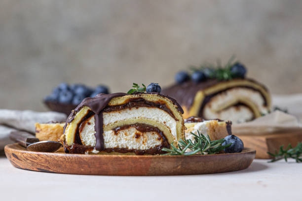 Sponge cake roll with chocolate and cream cheese decorated with chocolate glaze, blueberry and rosemary on parchment, light background. Biscuit swiss roll.
