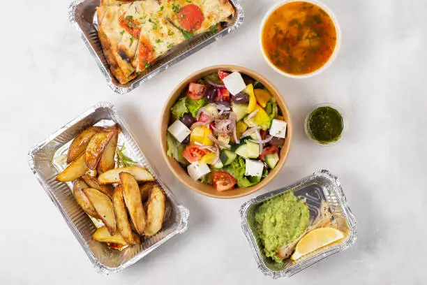Photo of Food delivery, italian cuisine: salad, focaccia, potatoes, minestrone soup, pesto sauce in boxes to take away. Vegetarian restaurant, simple and tasty business lunch. Top view on a white background