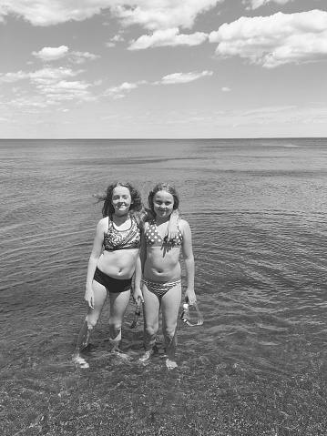 Two young girls, with their arms around each other, and standing near the shore of Lake Michigan, playing at the beach. Horizon and skyline can be seen in the background. Taken in Marinette, Wisconsin. Black and white photograph.
