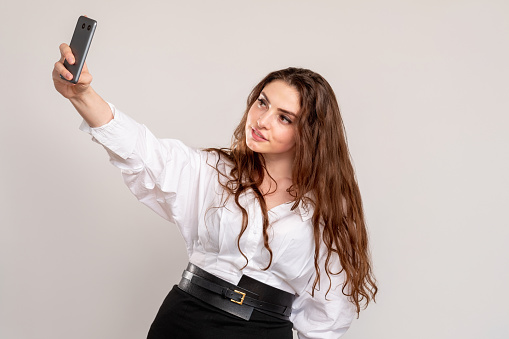 Business woman lifestyle. Mobile technology. Confident female leader in white shirt taking selfie on phone camera isolated on neutral empty space background. Social network. Online communication.