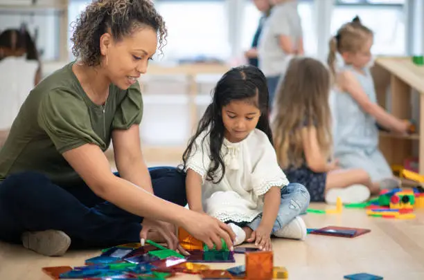 A female preschool teacher of African ethnicity helps one of her students build a tower using magnetic tiles.