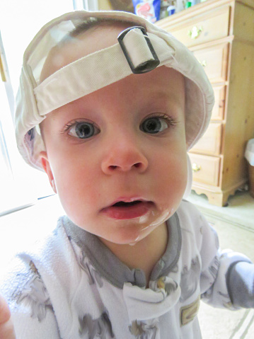 A wide angle close up of a young boy looking at the camera.  He is wearing a white baseball cap backwards. His face is one of curiosity and there is a bit of drool on his chin.