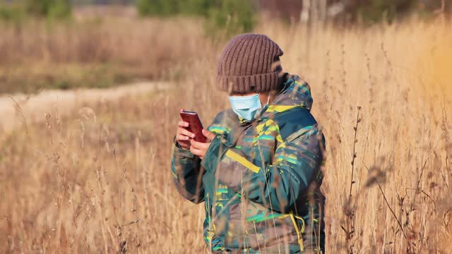 A boy in a medical mask the tall grass, he uses the Internet on his phone
