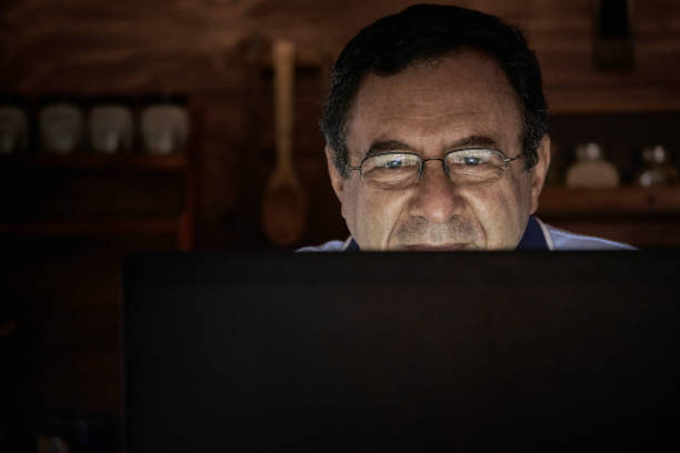 istock Senior Hispanic man using laptop in a domestic kitchen home office with low lighting 1296268335