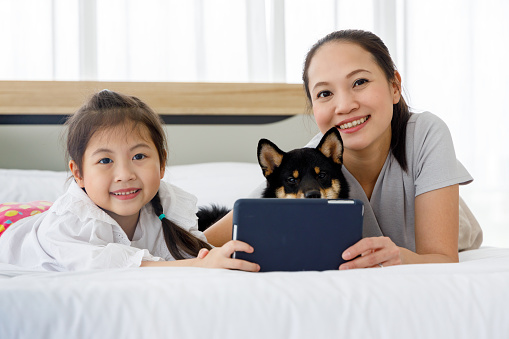 Happy Asian mother and daughter lying on bed and using digital tablet. Black Shiba Inu on bed between its owner. Concept family with dog at home.