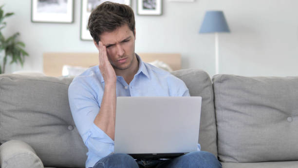 istock Handsome Young Man with Headache Working on Laptop while Sitting on Couch 1296271832