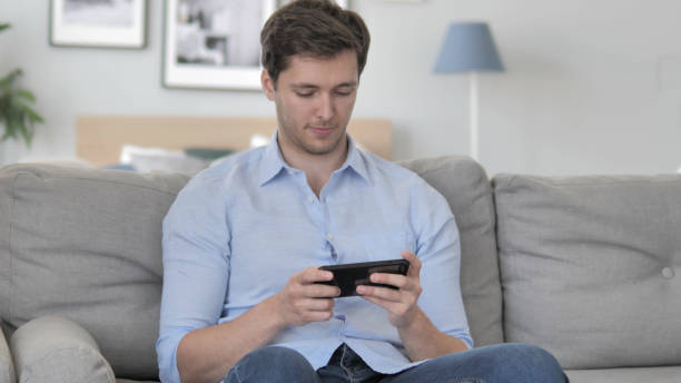 istock Young Man Playing Games on Smartphone, Sitting on Couch 1296271973