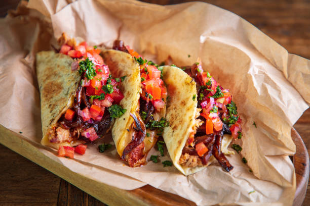 Serving of tacos with shredded roast pork and cheese filling on brown paper in wooden plate Serving of tacos with shredded roast pork and cheese filling, garnished with caramelized spanish onion and chopped tomato salsa sauce tacos stock pictures, royalty-free photos & images