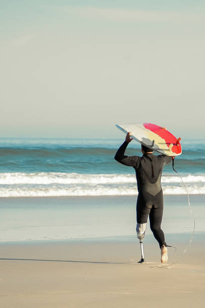 back view of handicapped surfer going to sea with board - surfing sport extreme sports success imagens e fotografias de stock