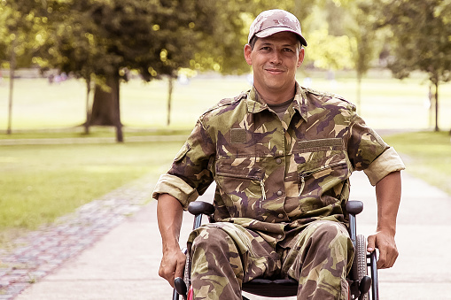 Happy disabled military man in wheelchair wearing camouflage uniform, moving on footpath in city park. Front view. Veteran of war or disability concept