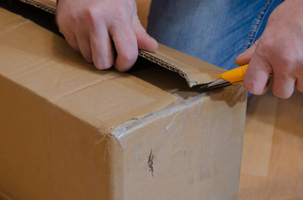 istock Man with yellow knife opens parcel in closed rectangular cardboard box on the floor. 1296289089