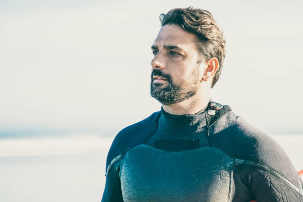 Caucasian athlete in black wetsuit holding surfboard and looking away. Portrait of serious attractive brunette surfer standing at coast near sea. Lifestyle, vacation and summer activity concept
