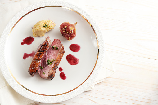 Slices of roasted duck breast with fig,  mashed parsnip and red cassis sauce on a white plate and a light wooden table, festive holiday dinner, copy space, high angle view from above, selected focus
