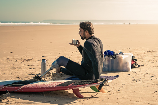 Caucasian surfer with artificial leg sitting and drinking tea from vacuum flask. Handsome brunette man relaxing on sand beach after surfing. Physical disability, lifestyle and extreme sport concept