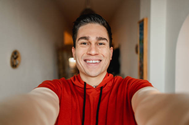 Young casual man smiling while  taking a selfie portrait looking at the camera inside the house.