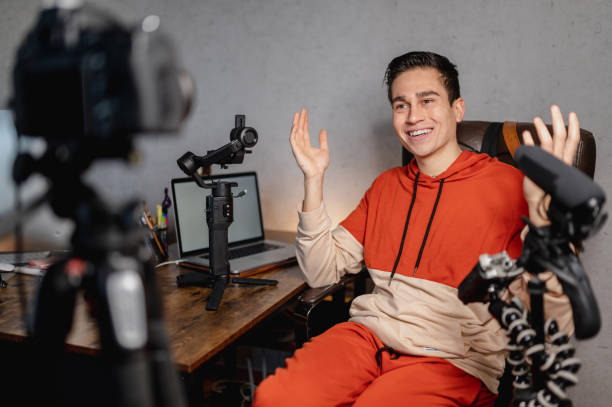 istock Young man sitting at the desk talking to the camera recording a tutorial video. Guy recording a video with professional equipments. 1296299499