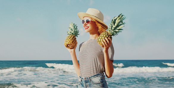 Portrait of happy smiling young woman with pineapples on the beach over sea background at summer day