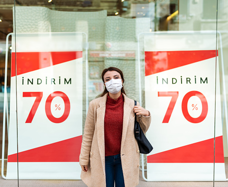 Beautiful girl wearing protective medical mask and fashionable clothes shows sale,discount clothes store. New normal lifestyle concept.