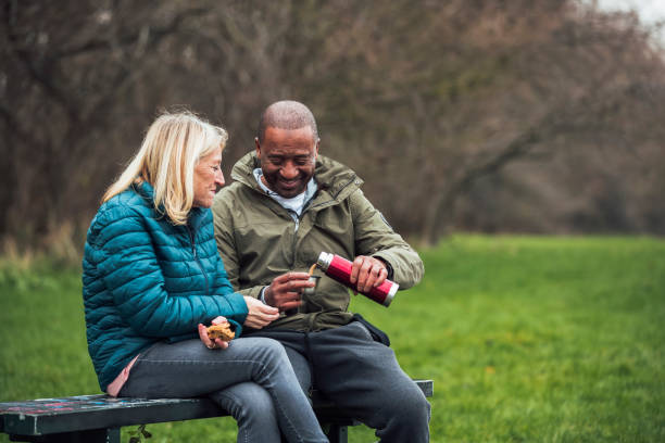 Senior Couple Picnic in the Park A senior mixed race couple wearing warm clothing and sitting on a park bench on a day in December. They are having a snack and a warm drink out of a flask. common couple men outdoors stock pictures, royalty-free photos & images