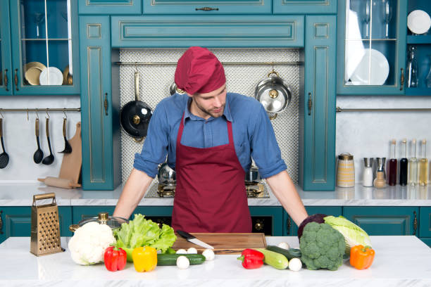 istock Cook in chef hat and apron at kitchen table. Cook man with vegetables and tools ready for cooking dishes. 1296310551