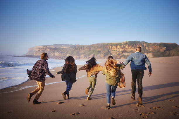 Friends group running from wave at the beach Back-view of multi-ethnic friends group running from wave at the beach during sunset runaway stock pictures, royalty-free photos & images