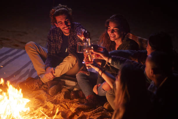 group of friends toasting drinks on beach bonfire at night - friendly fire imagens e fotografias de stock