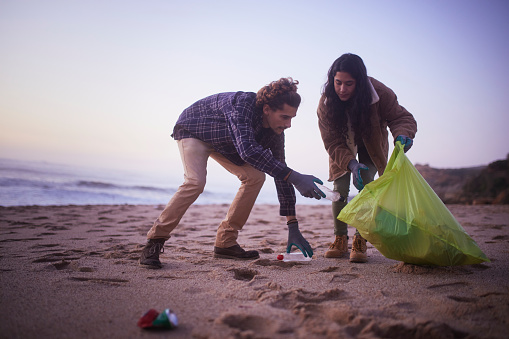 Two young friends picking up plastic trash on the beach during sunset