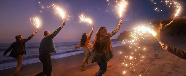 Back-view of group of young friends running with sparklers on the beach at night