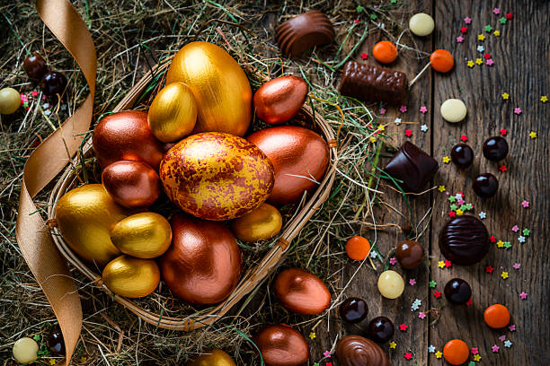 Holidays: hand painted gold and bronze colored Easter eggs in a wicker basket shot from above on rustic wooden table. The composition includes chocolate candies, sugar sprinkles and a gold colored ribbon. Predominant color is brown. High resolution 42Mp studio digital capture taken with Sony A7rII and Sony FE 90mm f2.8 macro G OSS lens
