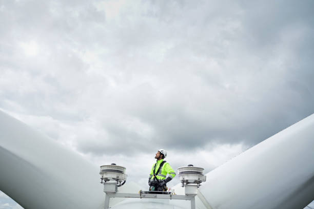 Professional rope access technician standing on roof (hub) of wind turbine between blades and antennas and looking up on the blade. Dramatic sky behind. Hands on hips stock photo