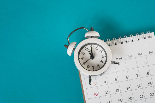 istock white analog alarm clock on blurred calendar with grunge blue background with copy space, business meeting schedule, travel planning or project milestone and reminder   concept ,panorama 1296319972