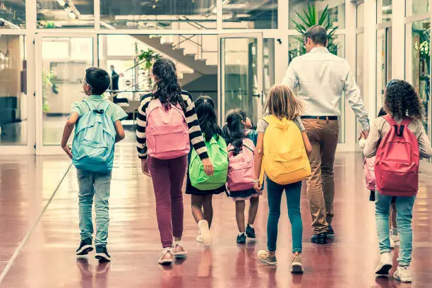 Schoolkids with colorful backpacks and male teacher walking through school hallway. Back view, full length. Education or back to school concept