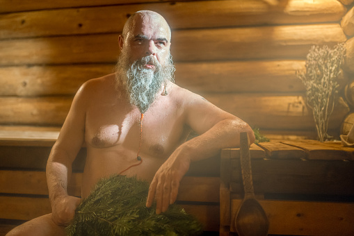The mature bearded man is relaxing at Russian steam bath (banya). He is sitting and looking at the camera