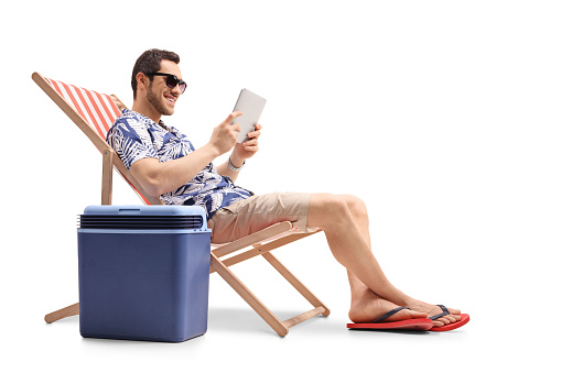 Young man sitting in a deck chair next to a cooling box and holding a digital tablet isolated on white background