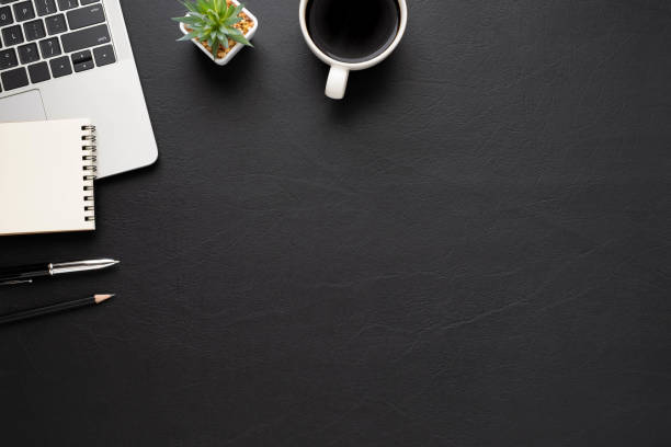 istock Top view above of Black leather office desk table with keyboard, notebook and coffee cup with equipment other office supplies. Business and finance concept. Workplace, Flat lay with blank copy space. 1296328026