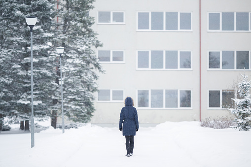 Young adult woman in warm coat walking on snow covered sidewalk in white winter day. Go to work or home. Back view.
