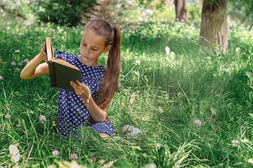 A school-age girl reading a book sitting on the grass in the park. Front view